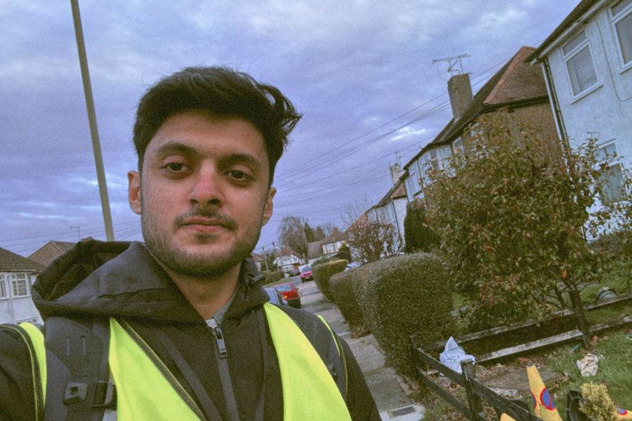 Muhammad in a hi-vis vest, taking a selfie on a residential street of front doors.