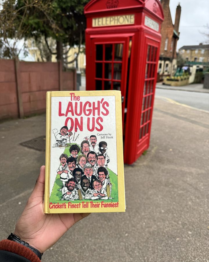 A cricket humour book, The Laugh's On Us, held up in front of a red phone box filled with books.